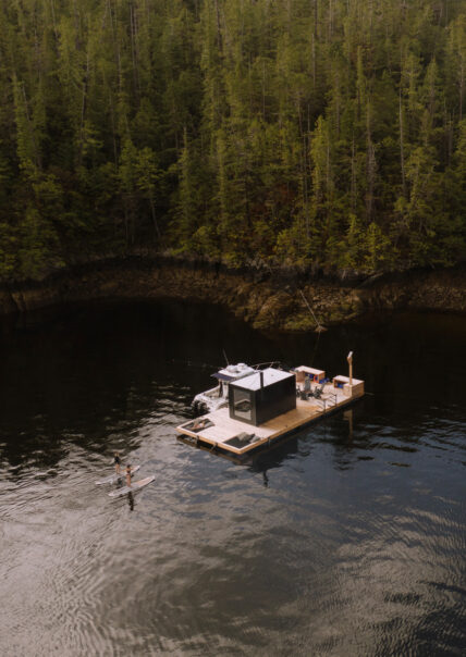 Aerial view of the floating sauna and dock with paddle boarders and guests lounging in remote Tofino