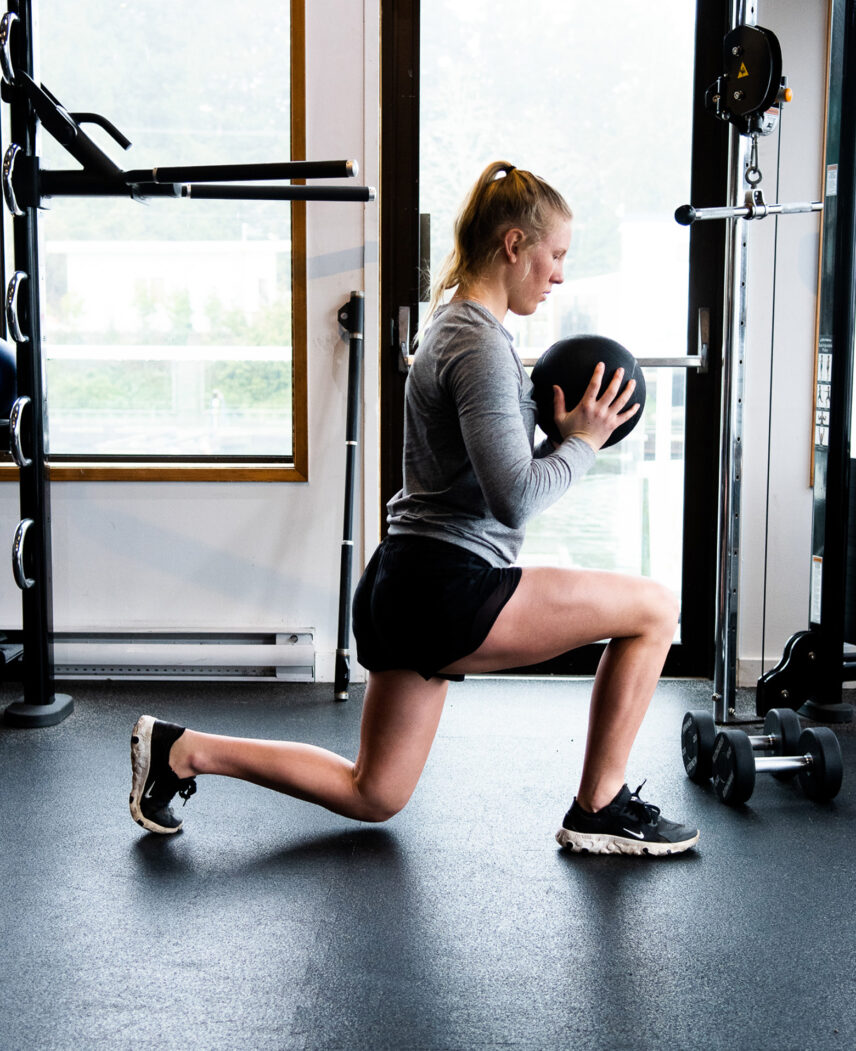 Woman performing a lunge with a medicine ball at Tuff Fit Gym, surrounded by natural light and fitness equipment