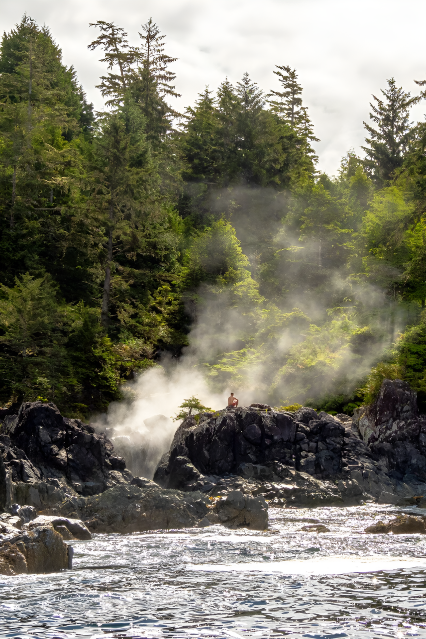 Steam rising from Hot Springs Cove pools nestled in rainforest, with a person resting on a coastal rock