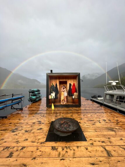 Two women at the entrance of the floating sauna with paddle boards, kayaks, and a rainbow overhead in Tofino