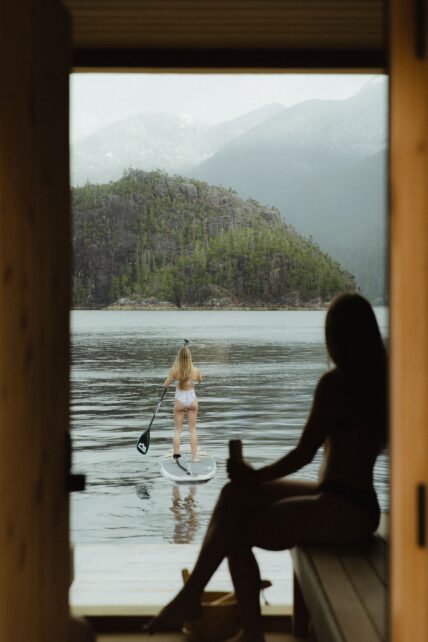 Silhouette of a woman inside the sauna watching another woman paddle board in Tofino’s remote inlet