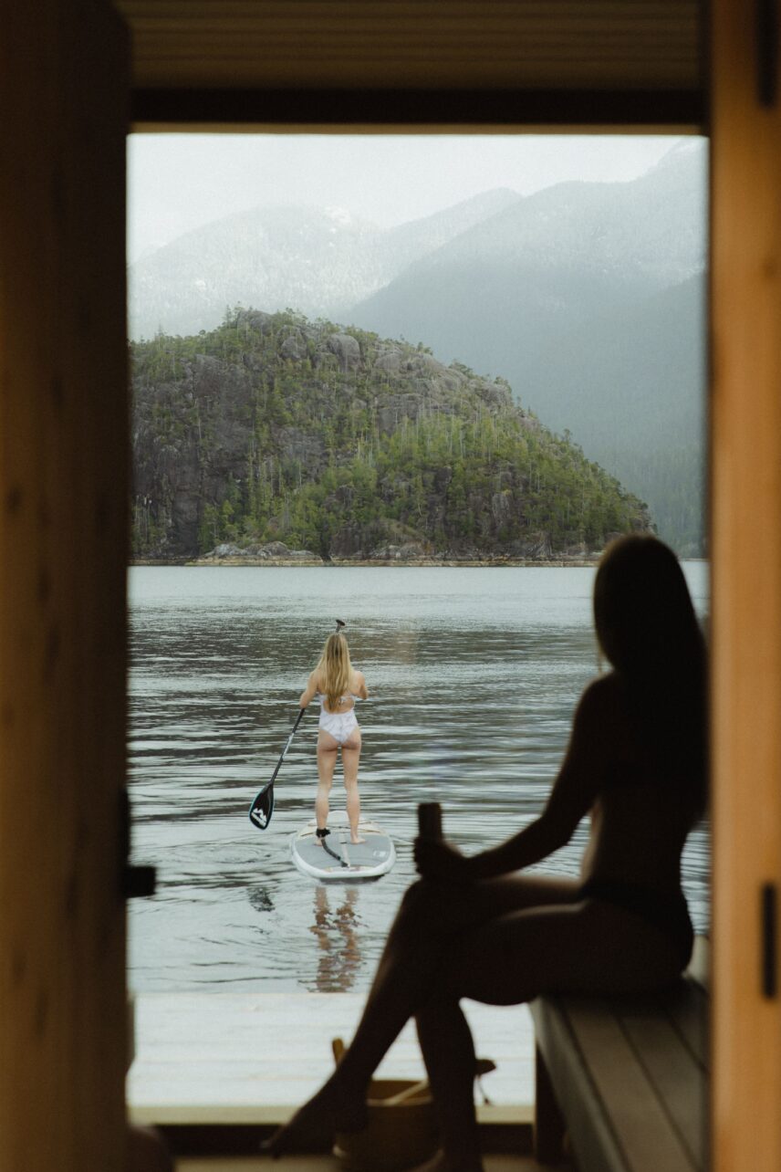 Silhouette of a woman inside the sauna watching another woman paddle board in Tofino’s remote inlet
