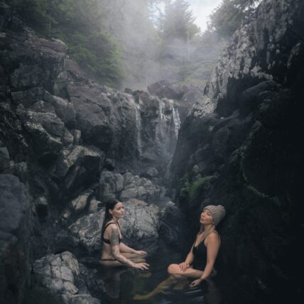 Two women soaking in natural hot spring pools with waterfalls and lush forest in the background