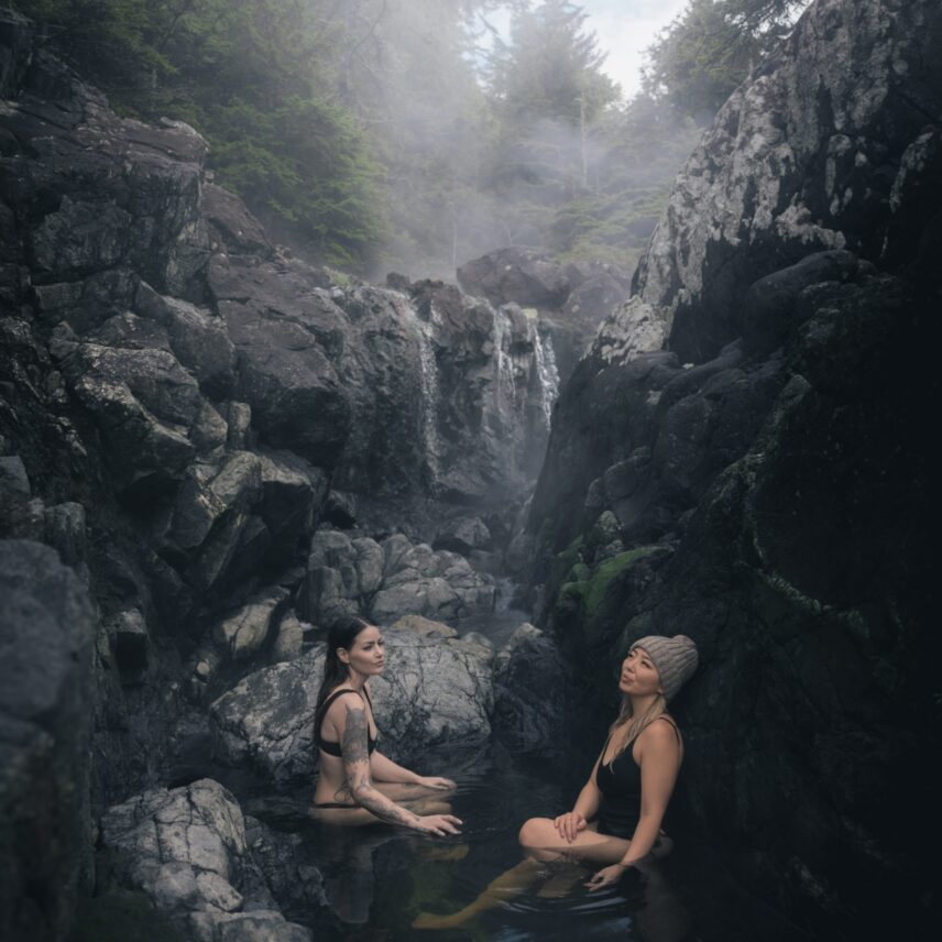 Two women soaking in natural hot spring pools with waterfalls and lush forest in the background