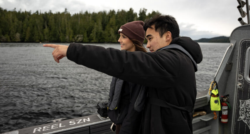 2 guests on a boat pointing and looking at the view under a gray winter sky